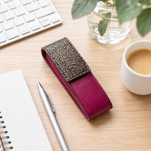 Op de afbeelding: Een bordeauxrode leren etui met een getextureerde, donkergekleurde bovenkant. De etui is open en onthult een contrasterend interieur. Een zilveren pen en een notitieblok liggen in de buurt, wat wijst op een werkomgeving.