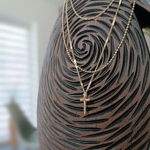Pode incluir: Um colar de corrente dourada em camadas com um pequeno pingente de cruz dourada. O colar é exibido em uma superfície de madeira marrom.