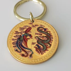 Può includere: Portachiavi color oro con un ciondolo rotondo con un design di drago e fenice. Il ciondolo ha un bordo dorato e un portachiavi dorato. Il drago è verde, rosso e oro, mentre la fenice è rossa, blu e oro.