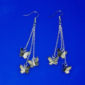 Könnte beinhalten: Silberne Ohrringe mit drei Schmetterlingsanhängern an jedem Ohrring. Die Schmetterlinge sind silberfarben mit schwarzen Akzenten.