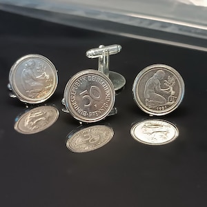 Könnte beinhalten: Ein Paar silberne Manschettenknöpfe mit einer deutschen 50-Pfennig-Münze aus dem Jahr 1981, auf der eine Frau einen Baum pflanzt. Die Manschettenknöpfe befinden sich auf einer schwarzen Oberfläche.