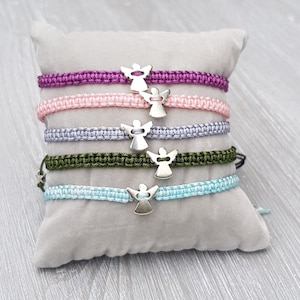 Könnte beinhalten: Fünf bunte Freundschaftsarmbänder mit silbernen Engelsanhängern. Die Armbänder liegen übereinander auf einem grauen Kissen. Die Armbänder sind rosa, lila, hellblau, grau und grün.