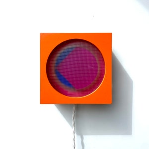 Puede incluir: Una lámpara de pared cuadrada naranja con un recorte circular que revela un diseño geométrico rosa, azul y blanco.