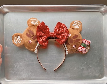 May include: Gingerbread-themed Minnie Mouse ears with a red sequined bow and white candy cane accents. The ears are brown and have a white icing design.