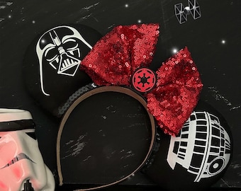 May include: Black mouse ears with a red bow and a white and black Star Wars character on each ear. The characters are Darth Vader and the Death Star.