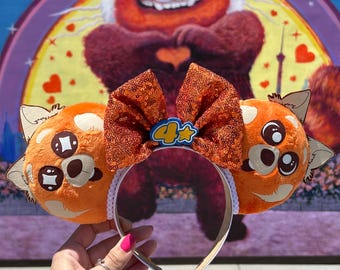 May include: Orange and brown plush red panda ears with a sparkly orange bow and a white headband. The bow has a 4+ charm in the center.
