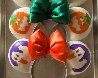 May include: Three pairs of Halloween-themed Minnie Mouse ears with different colored bows and character designs. The first pair has a black bow and a black cat design. The second pair has a green bow and a pumpkin design. The third pair has an orange bow and a ghost design.