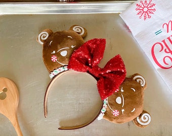 May include: A brown headband featuring two plush gingerbread bear ears with white icing details and a red sequin bow. The headband has a brown band and is decorated with candy cane accents. Festive holiday accessory.