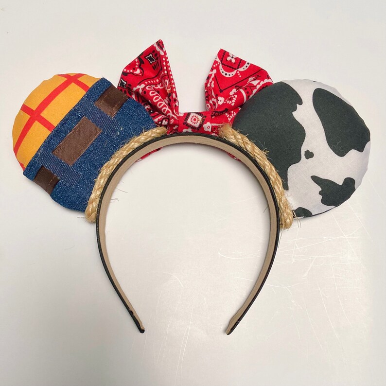 Peut inclure: Une paire d'oreilles de souris en tissu rouge, blanc et bleu avec une oreille en tissu denim et jaune d'un c&ocirc;t&eacute; et une oreille en tissu imprim&eacute; vache noir et blanc de l'autre. Les oreilles sont attach&eacute;es &agrave; un bandeau marron avec une bordure en corde.