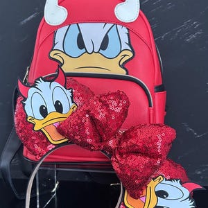 May include: Red backpack and headband set featuring a cartoon duck character with devil horns. The backpack has a matching character design. The headband has a large, sequined bow.