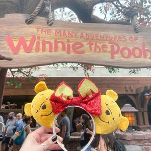 May include: Yellow Winnie the Pooh character mouse ears with a red sequined bow. The ears are plush and have a smiling Pooh face on each ear. The sign "The Many Adventures of Winnie the Pooh" is visible in the background.
