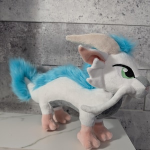 May include: A plush toy creature with white fur, blue accents, and pink feet. The toy has a long tail and a mane of blue fur. It has a gray snout, beige horns, and a green eye. The toy is standing on a white surface.