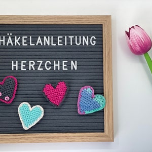 Könnte beinhalten: Eine schwarze Buchstabenwand mit dem Text "HÄKELANLEITUNG HERZCHEN" und vier gehäkelten Herzen in verschiedenen Farben: grau, pink, blau und türkis.