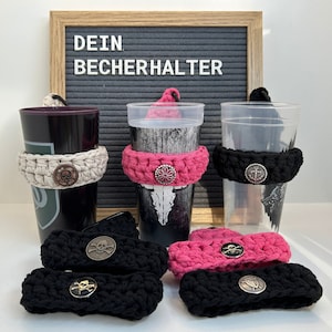 Könnte beinhalten: Drei gehäkelte Becherhalter mit Metallknöpfen in Form von Totenkopf und Anker. Die Becherhalter sind in beige, pink und schwarz. Das Schild darüber lautet "Dein Becherhalter".