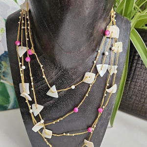Puede incluir: Un collar de múltiples hilos con cadenas finas de color dorado adornadas con pequeñas cuentas blancas, cuentas esféricas de color rosa vibrante y piezas planas de color blanco con forma de concha dispuestas en un diseño en cascada.