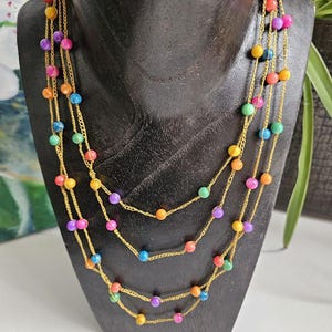 Può includere: Una collana a più fili con perline colorate nelle tonalità di rosso, arancione, giallo, verde, blu e viola. Le perline sono infilate su delicate catene color oro. La collana è esposta su un manichino nero.