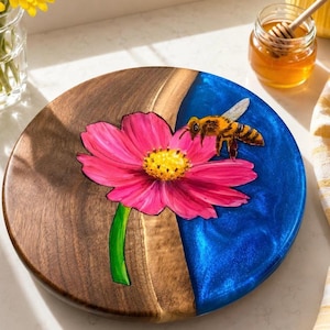 Può includere: Vassoio rotondo in legno con un design vivace. Metà del vassoio è in legno naturale, l'altra metà in resina blu lucida. Un grande fiore rosa con centro giallo e un'ape sono dipinti sul vassoio.