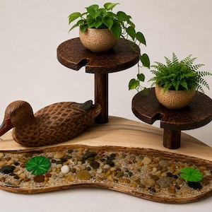 May include: A wooden display stand featuring a carved duck figure, two elevated circular platforms, and a base filled with pebbles and sand. Two textured pots containing green plants sit on the platforms, with two green lily pads on the base.