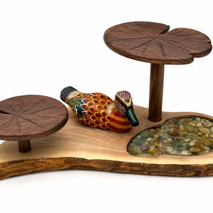 May include: A wooden display stand featuring a natural live-edge base with a small embedded resin pond filled with pebbles. A colourful carved wooden duck sits on the base next to two elevated dark wood platforms shaped like lily pads.
