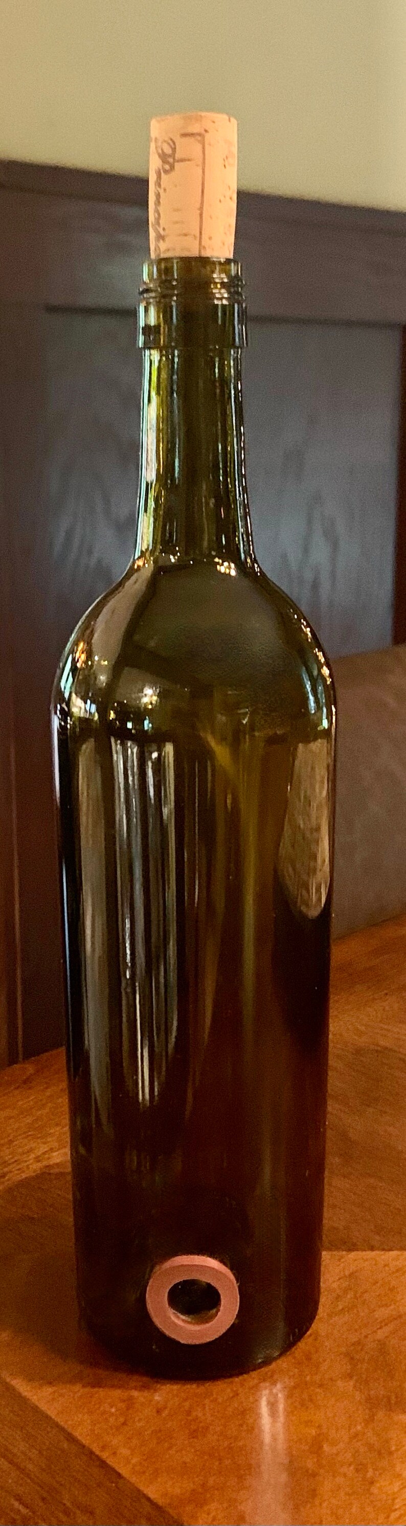 May include: A dark green glass bottle with a cork stopper. The bottle has a rounded body and a narrow neck. A copper-coloured ring is at the bottom. The cork has the word "Pinot" printed on it.