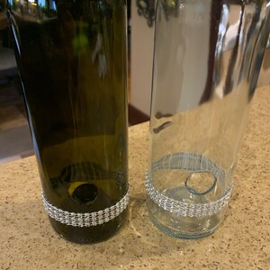 May include: Two upcycled wine bottles, one green and one clear, each featuring a decorative band of sparkling rhinestones near the base. The bottles have been cut and modified for a decorative or functional purpose.