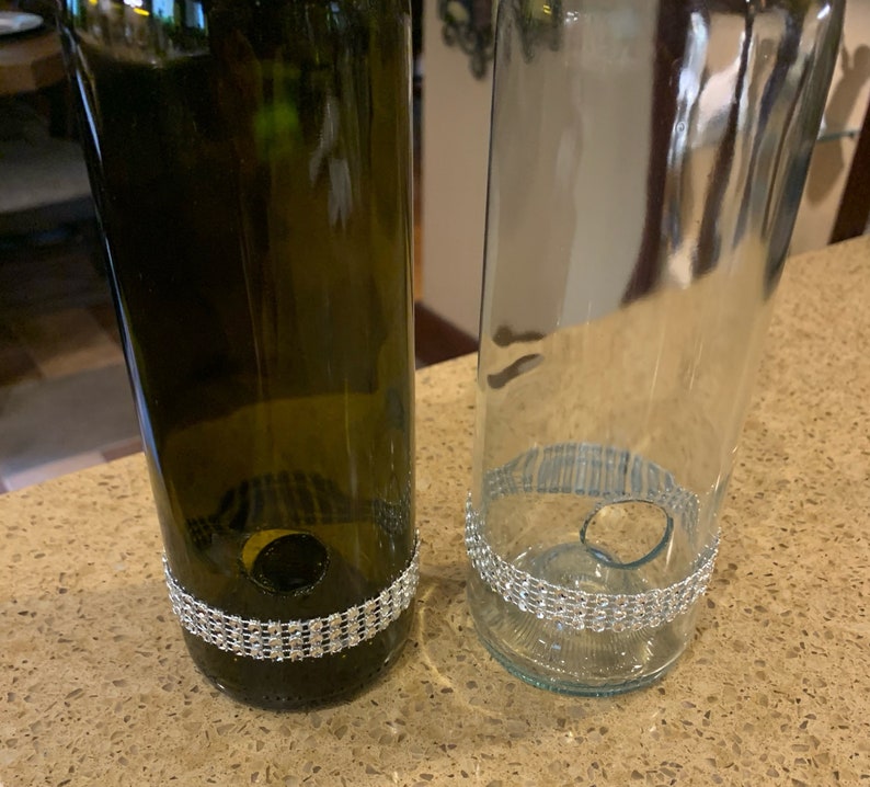 May include: Two upcycled wine bottles, one green and one clear, each featuring a decorative band of sparkling rhinestones near the base. The bottles have been cut and modified for a decorative or functional purpose.