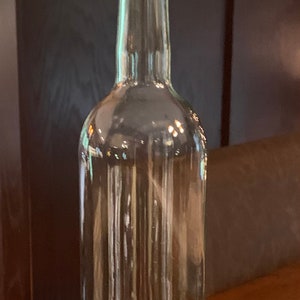 May include: A clear glass bottle with a cork stopper. The cork is labelled with the word "Domaine". The bottle has a white ring at the base. The bottle is empty and is likely for wine or other drinks.