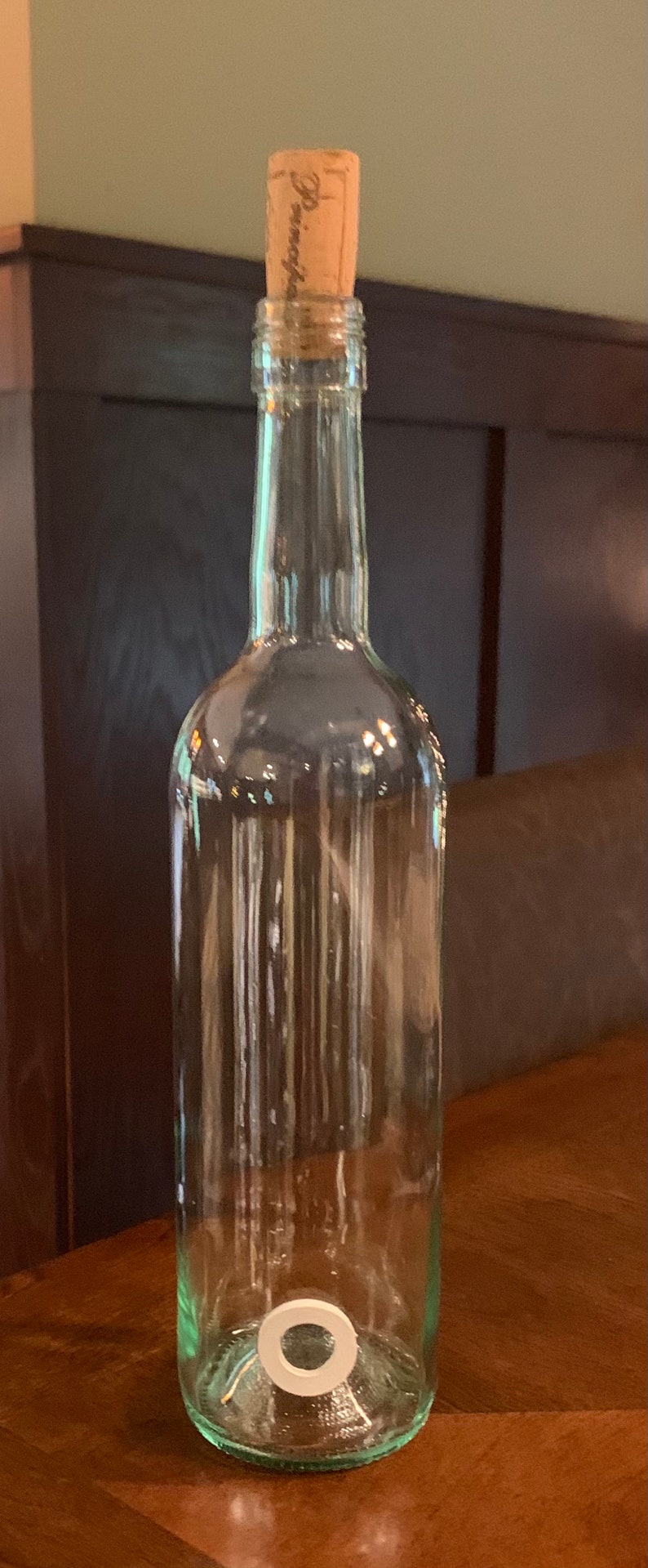 May include: A clear glass bottle with a cork stopper. The cork is labelled with the word "Domaine". The bottle has a white ring at the base. The bottle is empty and is likely for wine or other drinks.