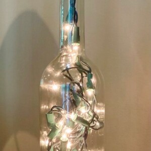 May include: A clear glass wine bottle repurposed as a decorative light. The bottle is filled with small, warm white string lights. A cork with text is at the top.