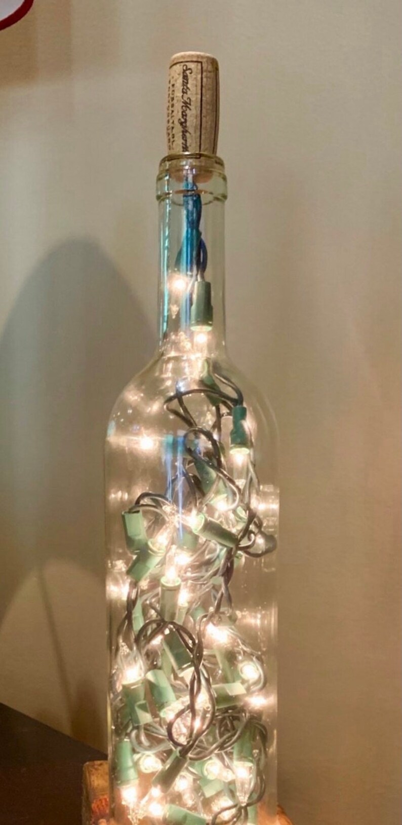 May include: A clear glass wine bottle repurposed as a decorative light. The bottle is filled with small, warm white string lights. A cork with text is at the top.