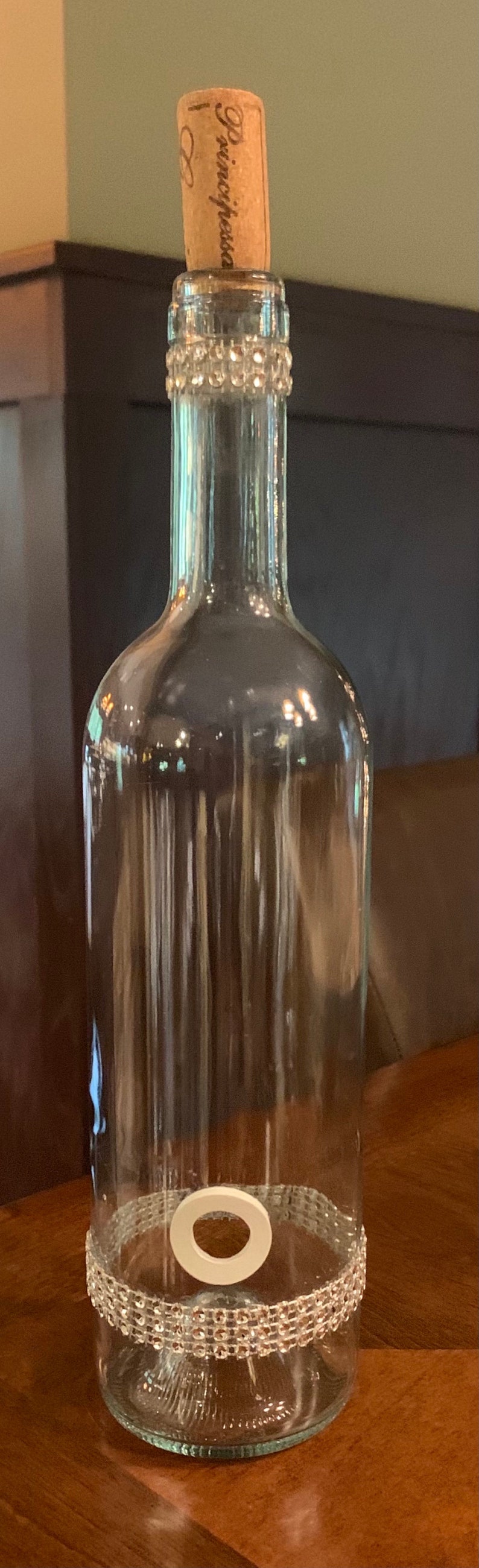 May include: A clear glass bottle with a cork stopper. The bottle is decorated with a band of clear rhinestones near the top and bottom. The cork has the word "Principessa" printed on it.