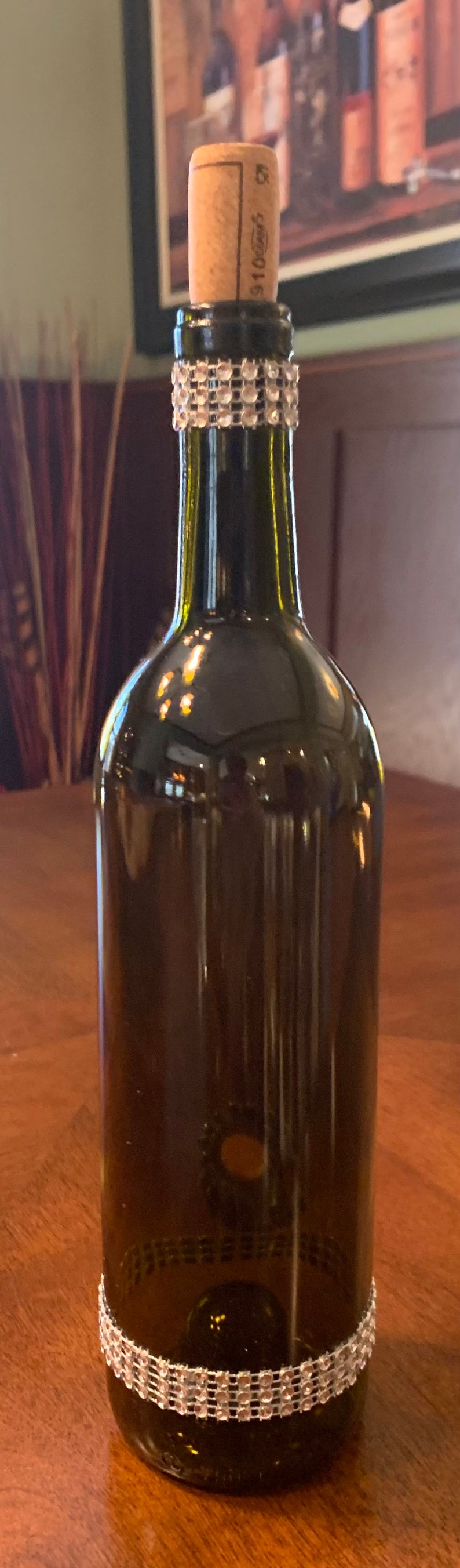 May include: A dark green glass bottle with a cork stopper. The bottle is decorated with two bands of clear rhinestones. The bottle is likely a decorative piece or a repurposed item.