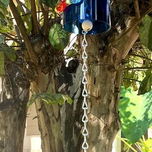 May include: A blue glass bottle wind chime suspended from a tree branch. The chime has colourful beads and a silver chain. The bottle is held by a wire and a cork. The background shows green leaves and a sunny day.