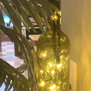 May include: A green glass bottle filled with small, warm white fairy lights. The bottle sits on a stone base, with green leafy branches in the foreground. The lights create a soft, ambient glow.