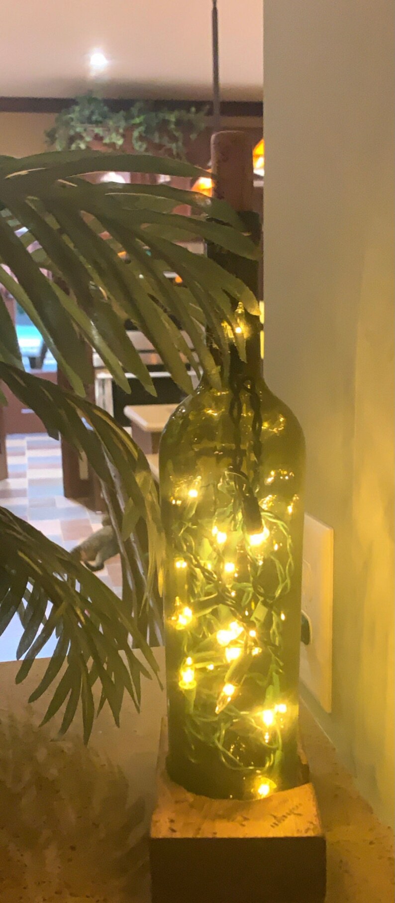May include: A green glass bottle filled with small, warm white fairy lights. The bottle sits on a stone base, with green leafy branches in the foreground. The lights create a soft, ambient glow.