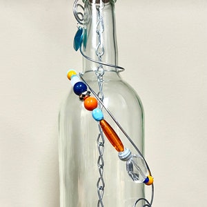 May include: A clear glass bottle with a decorative wire and bead embellishment. The bottle is topped with a cork and a metal hook. The wire design features colourful beads and a small charm. The cork reads "MAISON WINE".