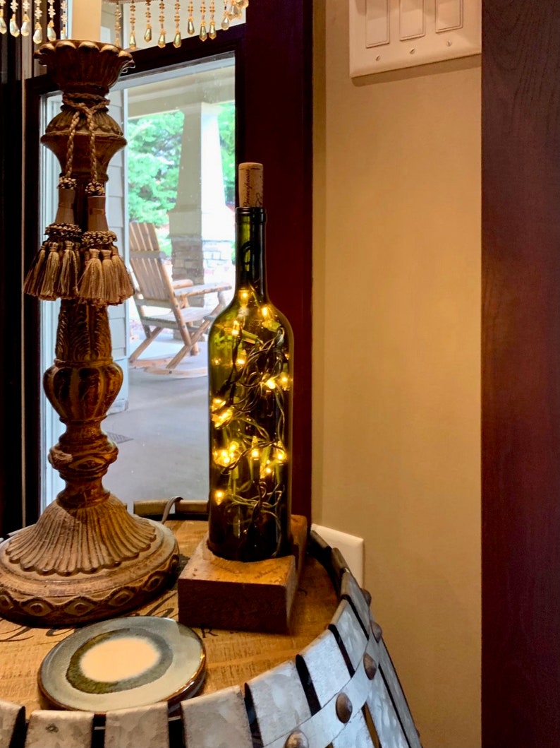 May include: A green wine bottle, repurposed as a decorative item, with a cork and fairy lights inside. The bottle is placed on a wooden block. The lights emit a warm glow. The bottle is displayed on a wooden barrel.