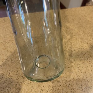 May include: Clear glass bottle with a rounded base and a circular opening. The bottle is cylindrical and appears to be empty. The glass has a slight green tint. The bottom of the bottle has embossed text.