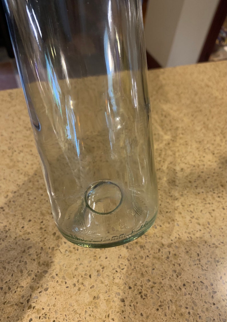 May include: Clear glass bottle with a rounded base and a circular opening. The bottle is cylindrical and appears to be empty. The glass has a slight green tint. The bottom of the bottle has embossed text.