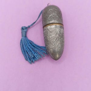 May include: A small, bullet-shaped metal container with a textured, engraved surface featuring swirling patterns. The two-piece object has a thin gold-toned band at the center and is attached to a bright blue decorative tassel made of twisted cord.