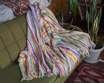 May include: A colorful striped crocheted throw blanket with fringe on a green couch. The blanket is made of various colors, including pink, blue, green, yellow, and purple.