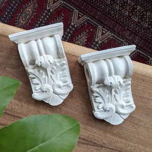 May include: Two white plaster corbels with ornate floral designs. The corbels are on a wooden surface.