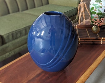 May include: A deep blue ceramic vase with a smooth, glossy finish and a swirling design. The vase is sitting on a wooden table.