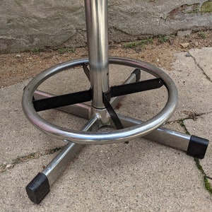 May include: Close-up of a chrome bar stool base with a black metal ring and a footrest. The stool has a swivel base and is designed for use in a bar or kitchen setting.