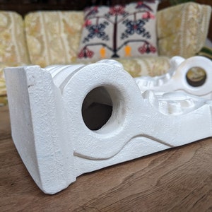 May include: Two white ceramic shelf brackets with intricate designs and a hole in the center. The brackets are on a wooden surface.