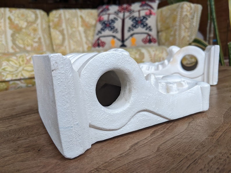 May include: Two white ceramic shelf brackets with intricate designs and a hole in the center. The brackets are on a wooden surface.