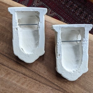 May include: Two white plaster wall sconces with a simple design. Each sconce has a rectangular opening with a metal bar across the top. The sconce on the left has the number "1176" inscribed on the front.
