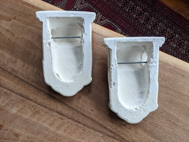 May include: Two white plaster wall sconces with a simple design. Each sconce has a rectangular opening with a metal bar across the top. The sconce on the left has the number "1176" inscribed on the front.