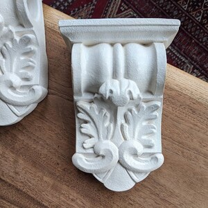 May include: Two white ornate corbels with a decorative floral design. The corbels are made of a lightweight material and have a smooth, painted finish.