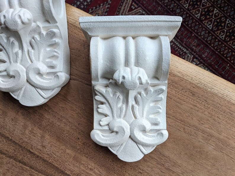 May include: Two white ornate corbels with a decorative floral design. The corbels are made of a lightweight material and have a smooth, painted finish.